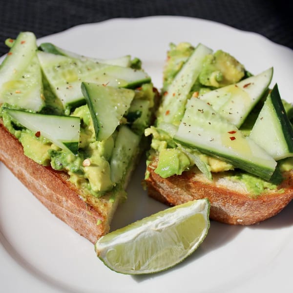 Avocado toast topped with sliced cucumbers, spices, and a lime wedge on a white plate.