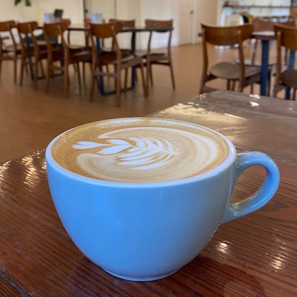 A light blue mug of latte with leaf art sits on a wooden table at The Riv Cafe.